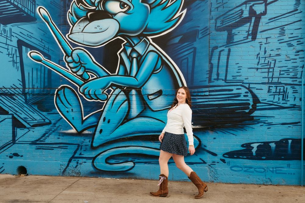 A person poses in front of a vibrant blue street art mural featuring a cartoon character in an urban setting.