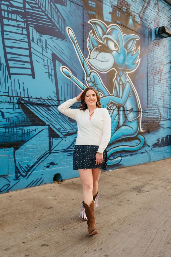 A person poses playfully in front of a blue street art mural featuring a cartoon character with a baseball bat.