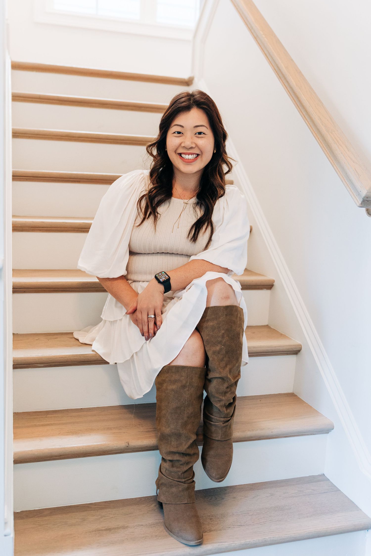 A person in brown suede boots sits casually on wooden stairs in a bright interior space, wearing a white top.