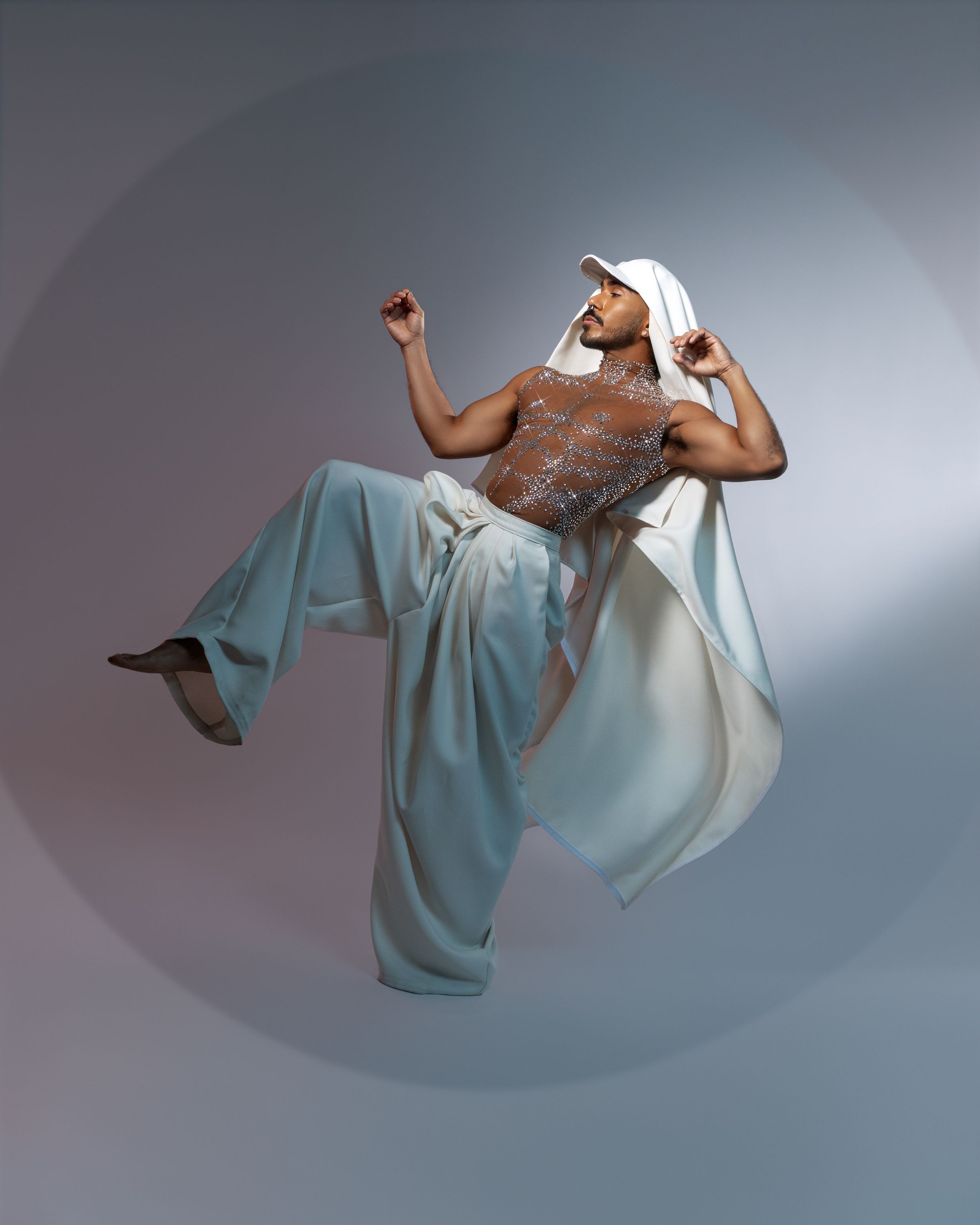 Athletic figure in white cap and flowing light blue pants performs dynamic mid-air dance pose against gray backdrop.