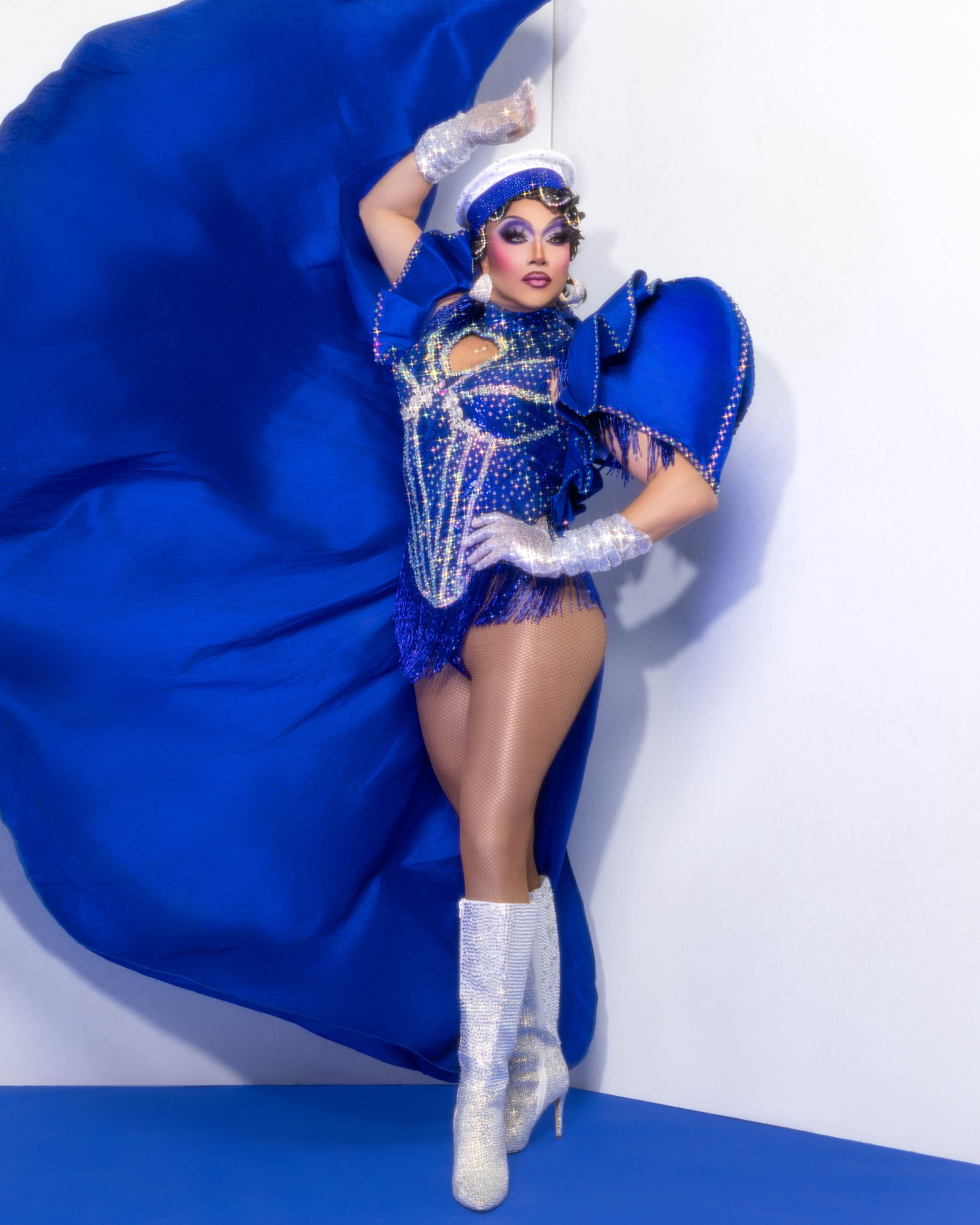 Drag performer in a dazzling royal blue rhinestone costume with white boots and flowing cape strikes a dramatic pose.