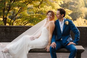 A couple in wedding attire sits together on a wooden bench surrounded by golden autumn foliage at sunset.