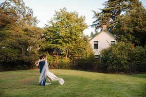 A couple in wedding attire dances and spins together on a grassy lawn during golden hour.
