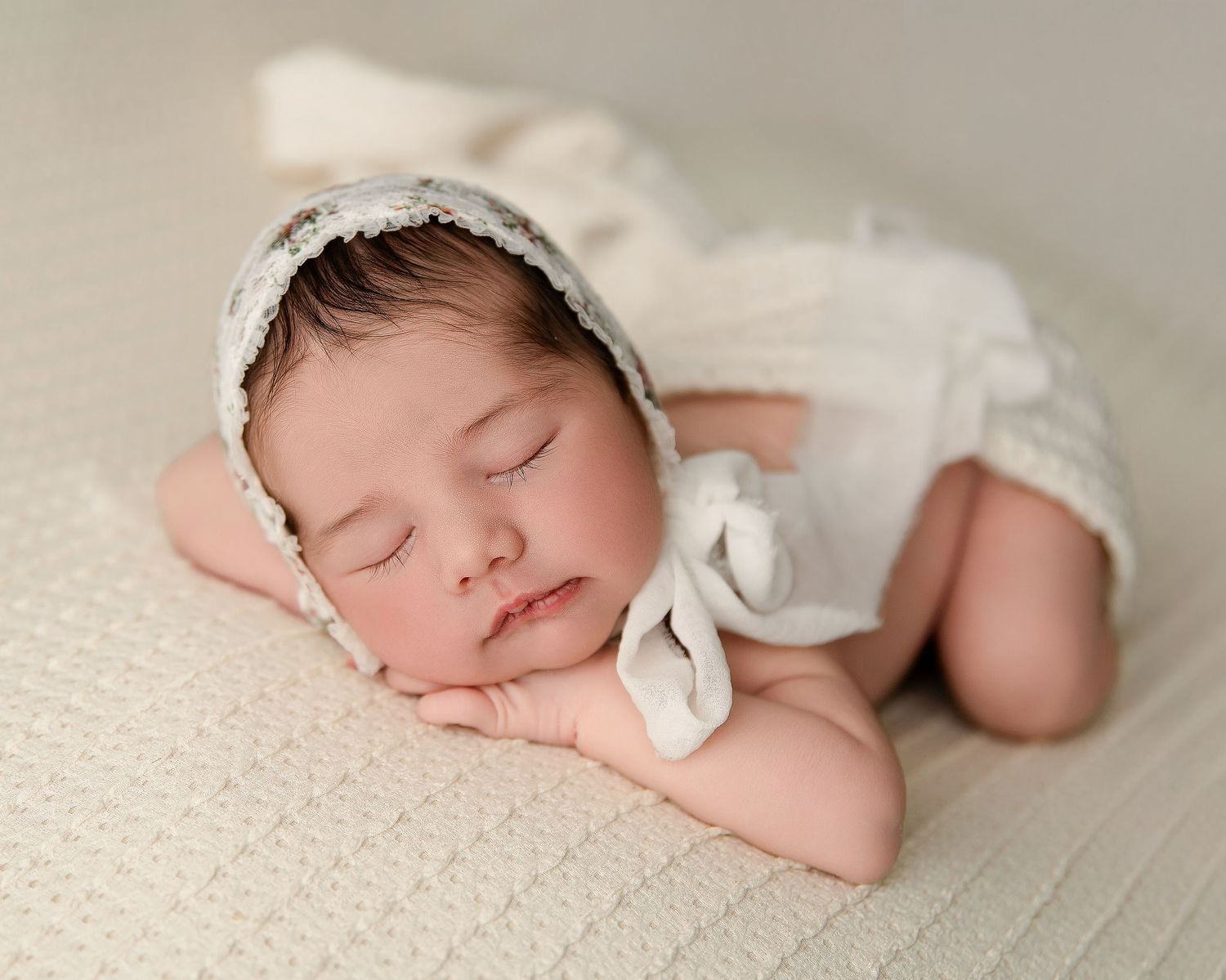 A sleeping newborn baby swaddled in white fabric lies peacefully on a soft cream-colored blanket.