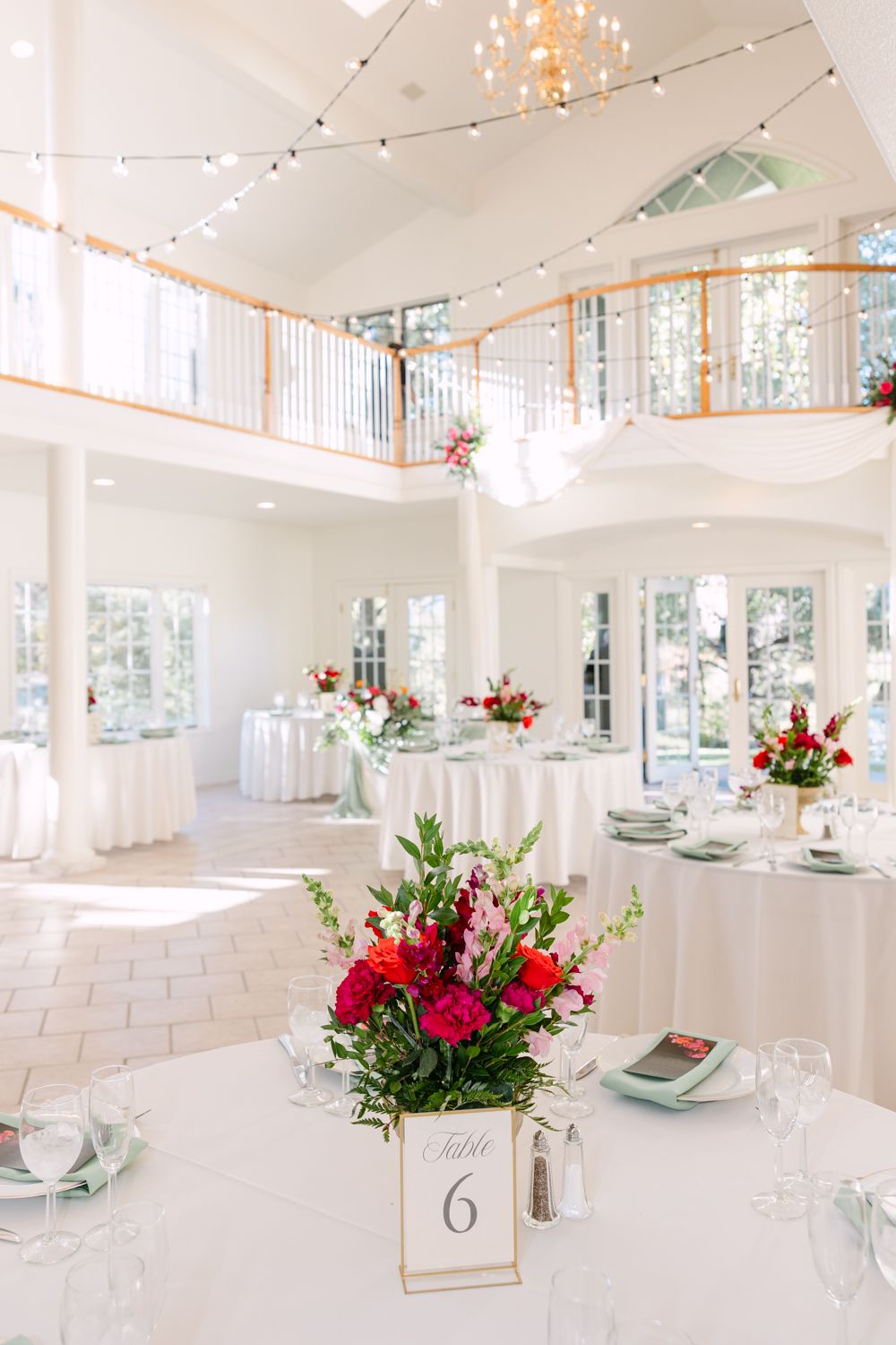 Elegant wedding reception venue with grand staircase, string lights, and round tables set with white linens and floral centerpieces.