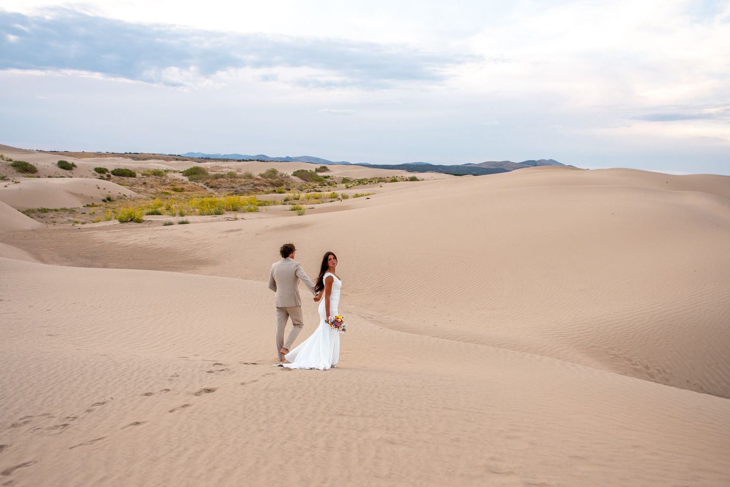 Two figures walk along desert sand dunes leaving footprints behind in the golden sand during sunset.