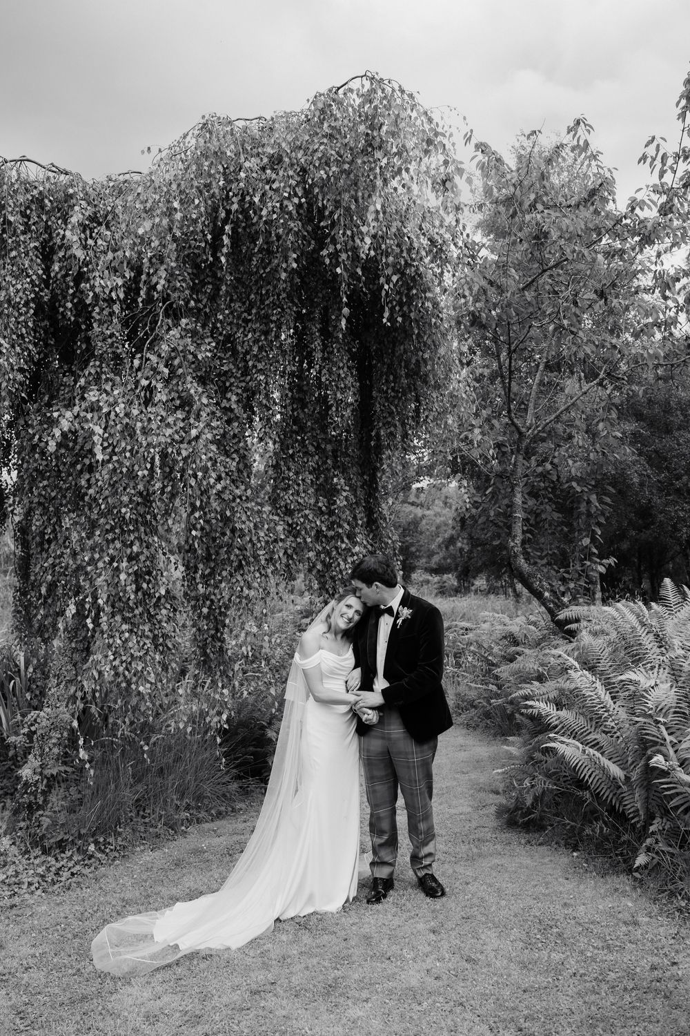 Black and white photo of a bride in a flowing gown and a groom in a dark suit embracing beneath a weeping tree in a garden.