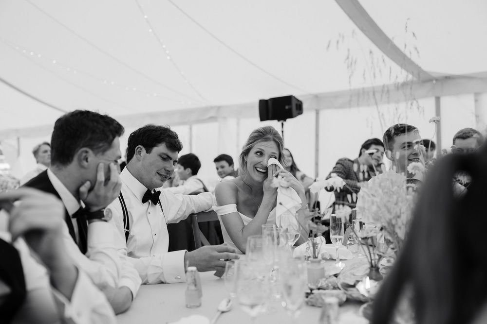 Emotional bride wipes away happy tears at a wedding reception table as groom and guests smile under a white marquee tent.