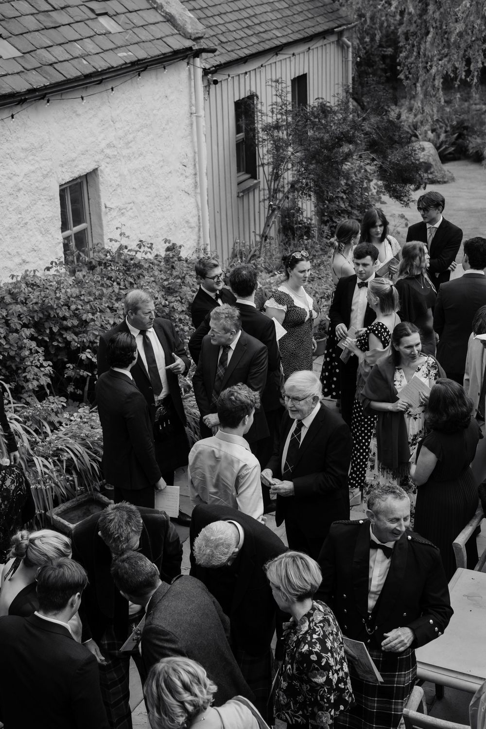 Black and white photo of formally dressed wedding guests mingling outdoors beside a stone cottage, with a man in a kilt in the foreground.