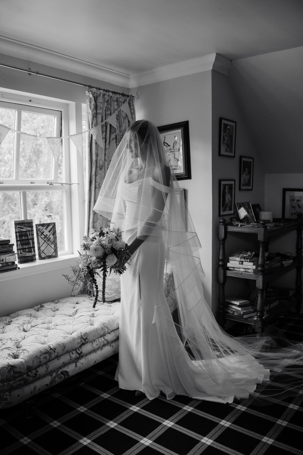 Black and white photo of a bride in a long veil and gown holding a bouquet beside a sunlit window in a cozy room with plaid carpet.