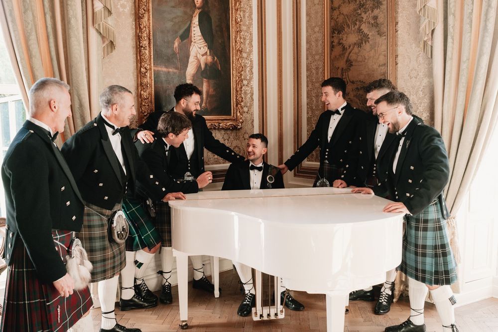 Groomsmen in traditional Scottish kilts and black jackets gather around a white grand piano in an elegant room with ornate paneling.