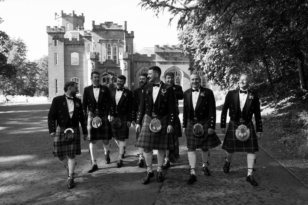 Groomsmen in traditional kilts, jackets and sporrans walk together along a tree-lined path in front of a Scottish castle in black and white.