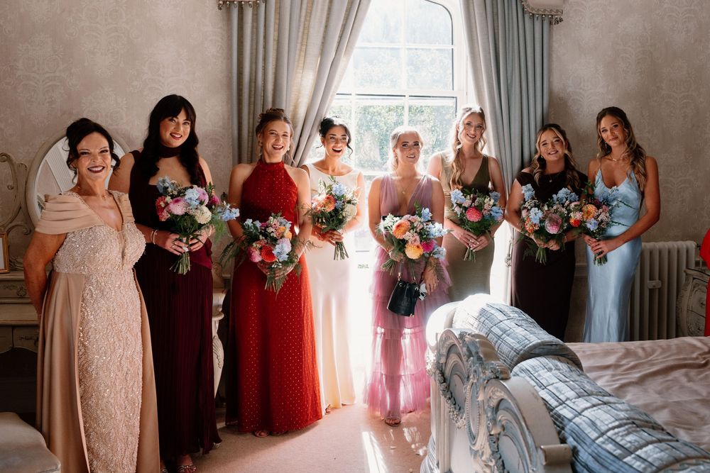 Bridesmaids in assorted dresses hold colorful bouquets while standing in a row beside a bright window in an elegant bridal suite.