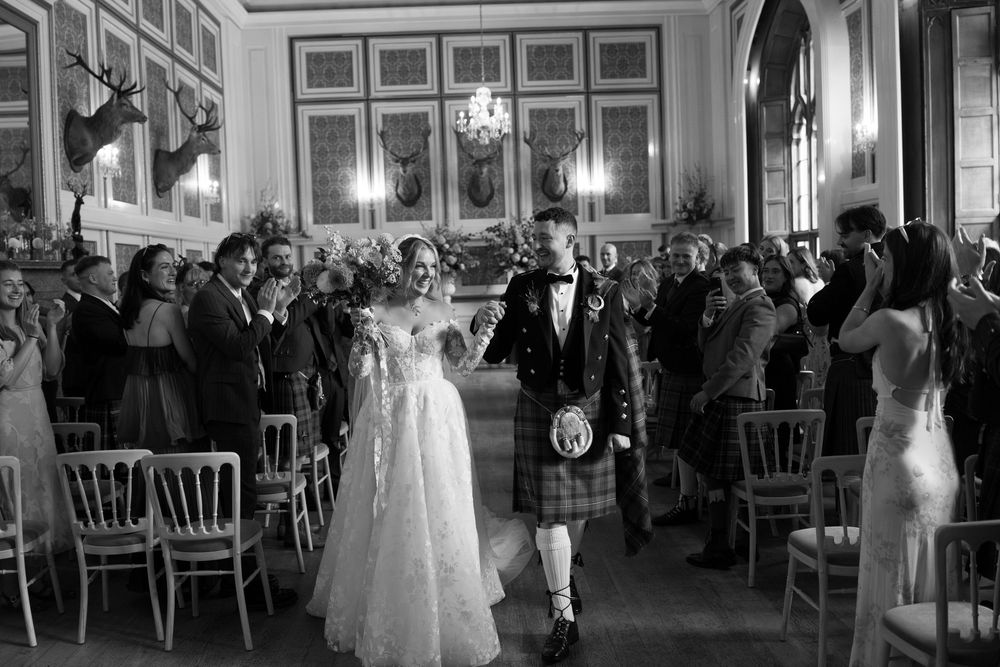 Bride in a lace gown and groom in a kilt walk hand in hand past seated wedding guests in a grand hall with mounted stag heads.