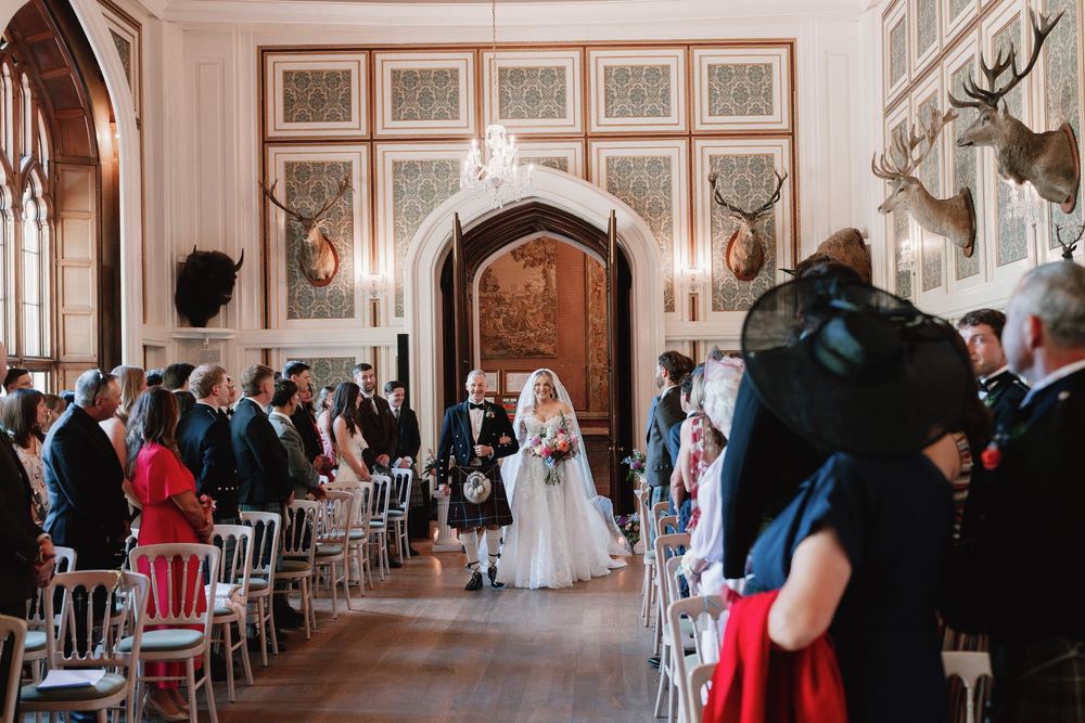 Bride in a white gown walks down the aisle on a man's arm as seated wedding guests look on in a grand hall decorated with mounted antlers.