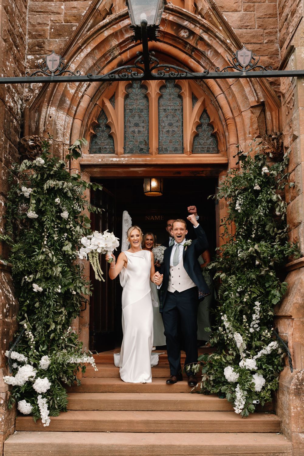 Joyful bride in a white gown and groom with raised fist exit a stone church doorway framed by lush white floral arrangements.