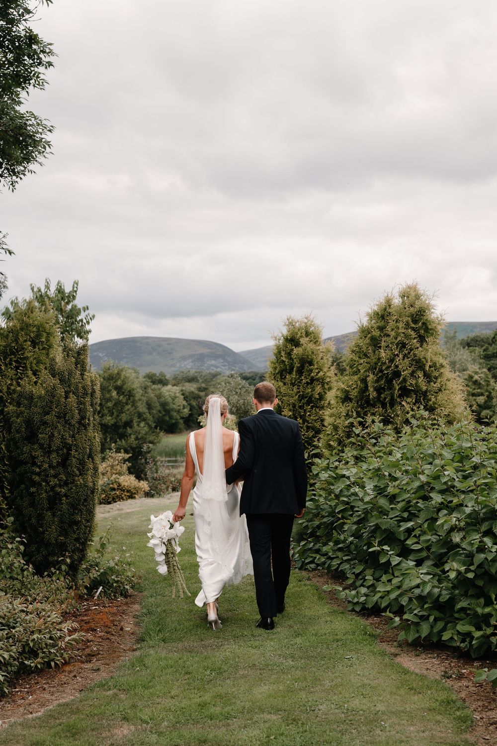 Bride in a long white gown and veil holding a bouquet walks hand in hand with a groom along a grassy garden path toward distant hills.