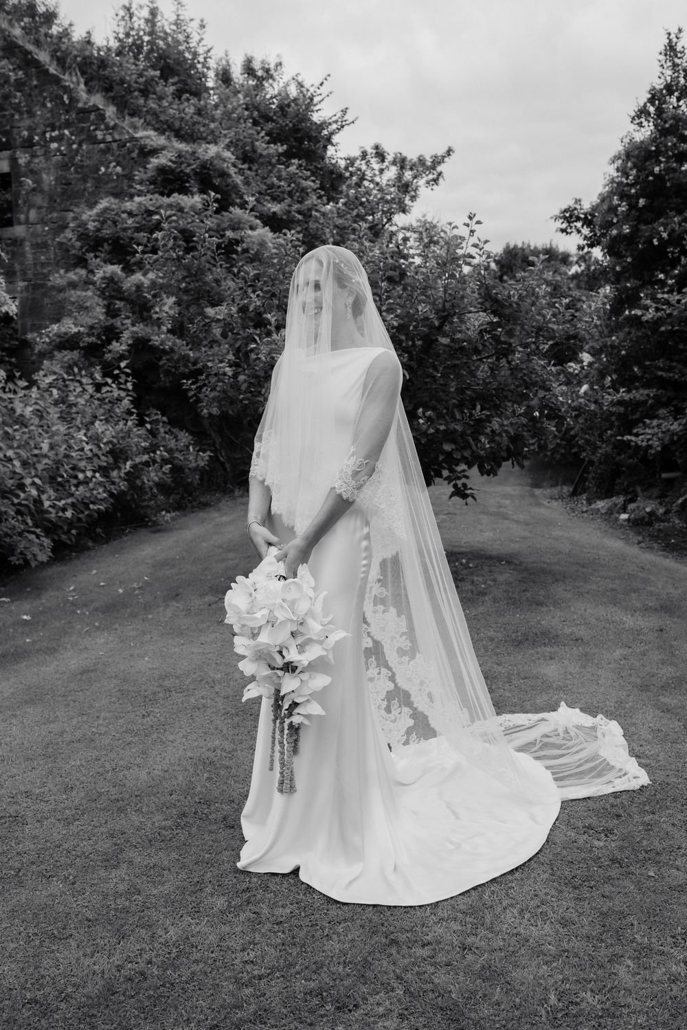 Black and white photo of a bride in an elegant gown and long lace-trimmed veil holding a cascading white bouquet in a lush garden.
