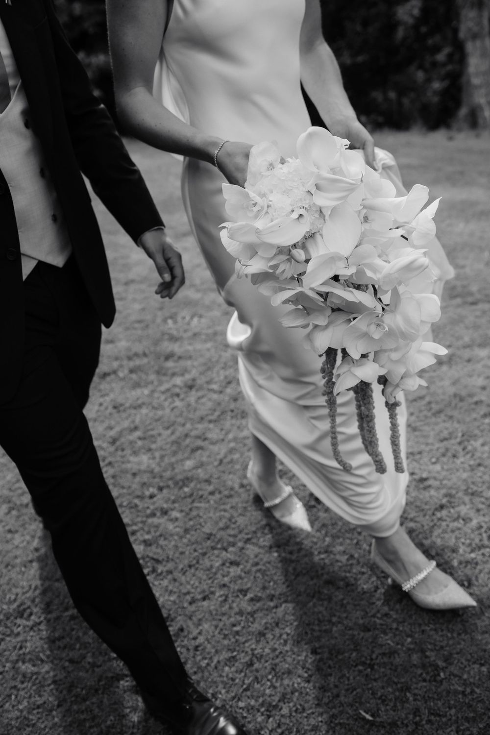 Black and white photo of a bride in a satin gown carrying a cascading white orchid bouquet while walking on grass beside a groom in a suit.