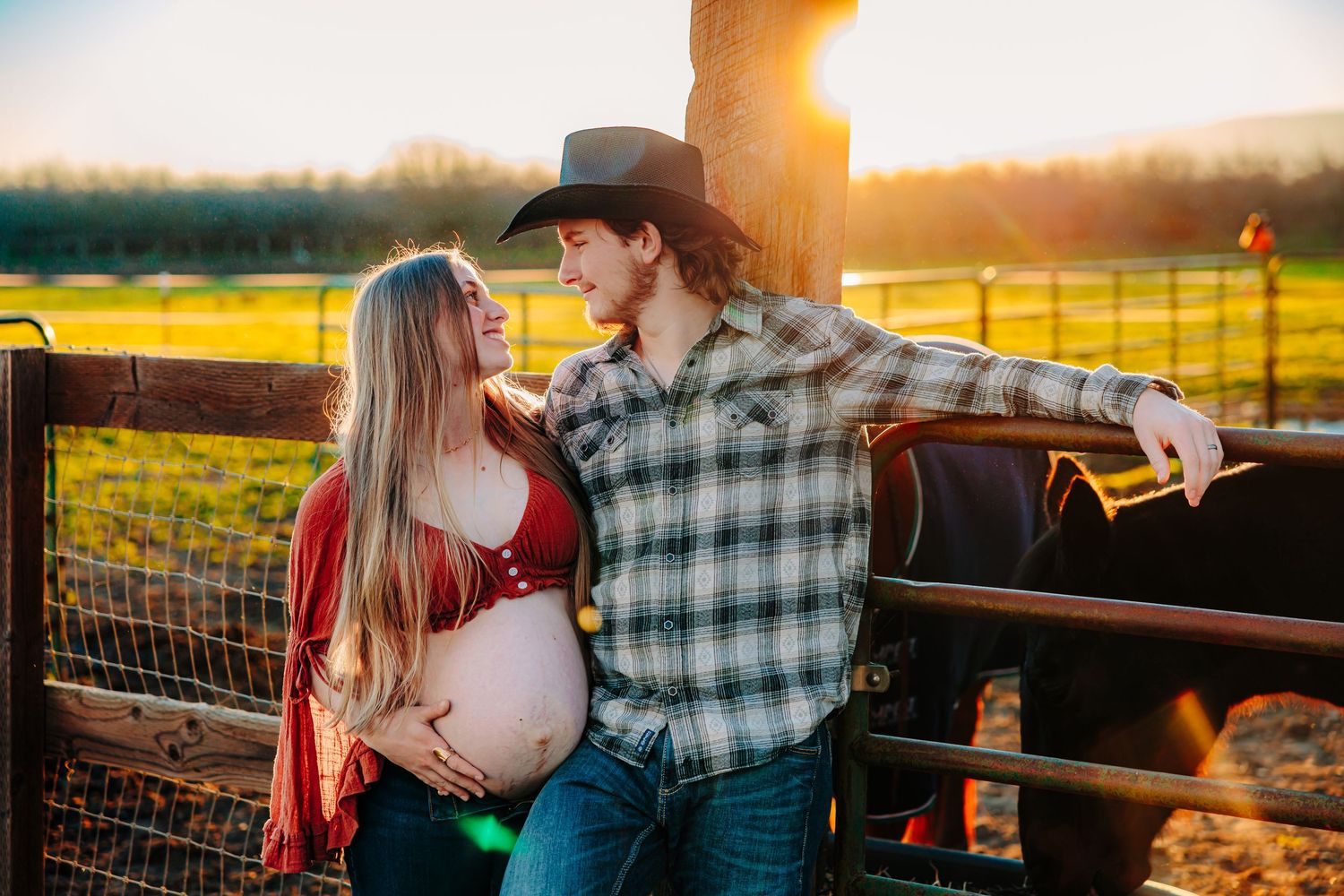 A couple shares a romantic moment at sunset by a wooden fence on a ranch, with warm golden light illuminating the scene.