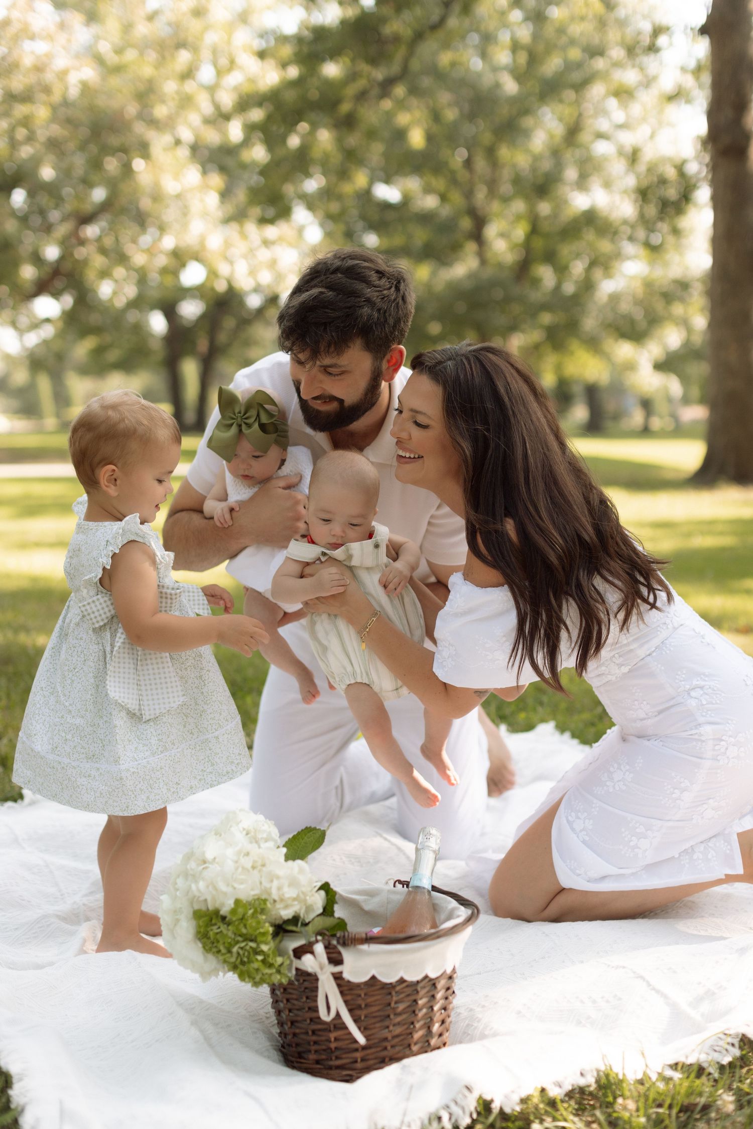 Family having a picnic together on a white blanket in a sunny park with a wicker basket.