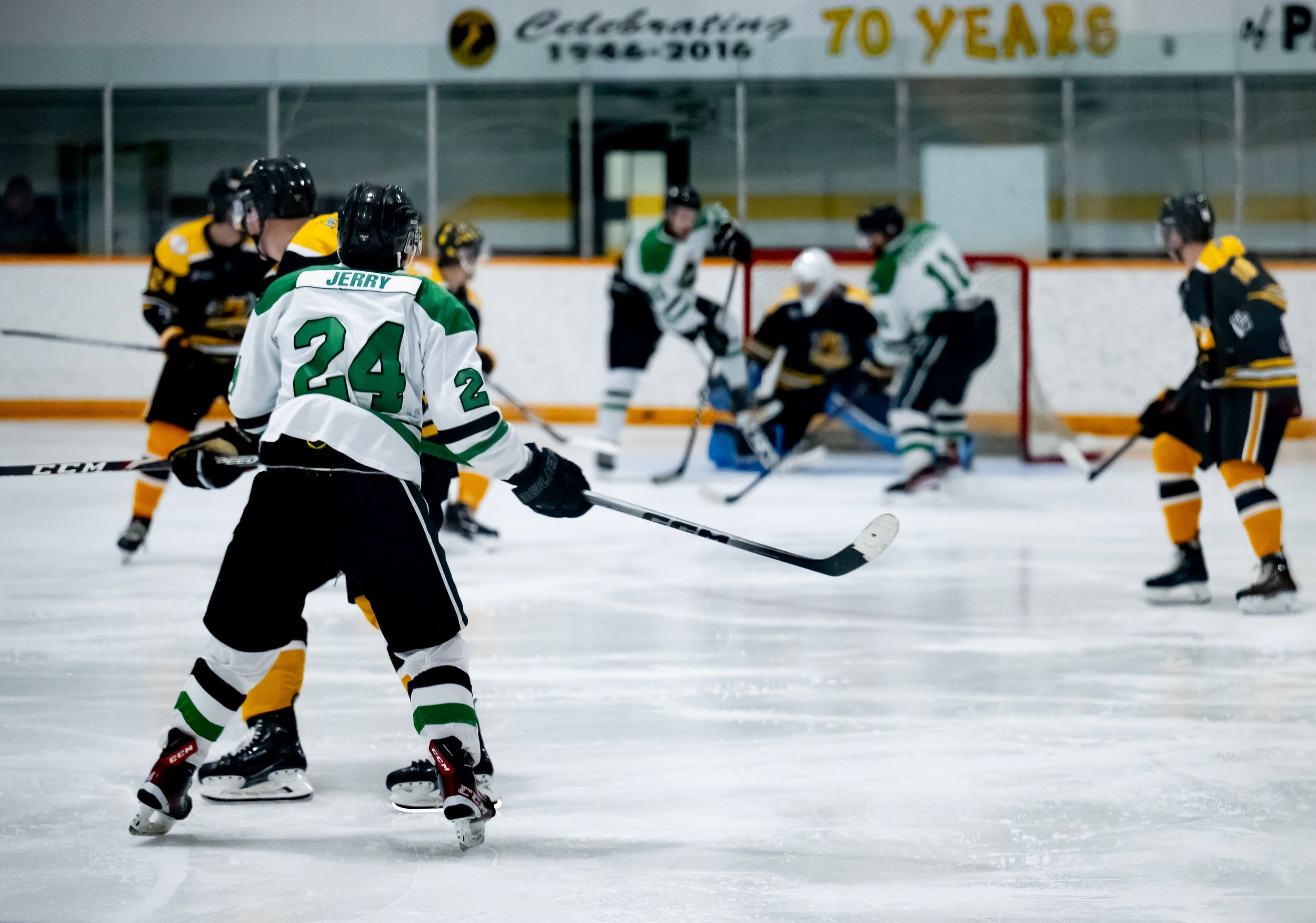 Hockey players in green and yellow jerseys compete during an ice hockey game with a 70th anniversary banner visible.