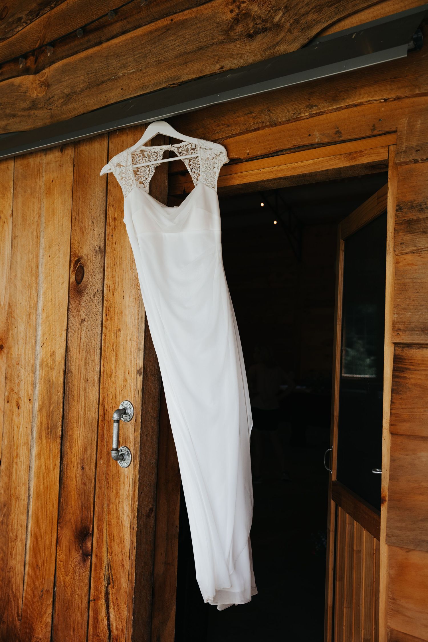 A sleek white wedding dress with lace details hangs on a wooden wall near a rustic doorway.
