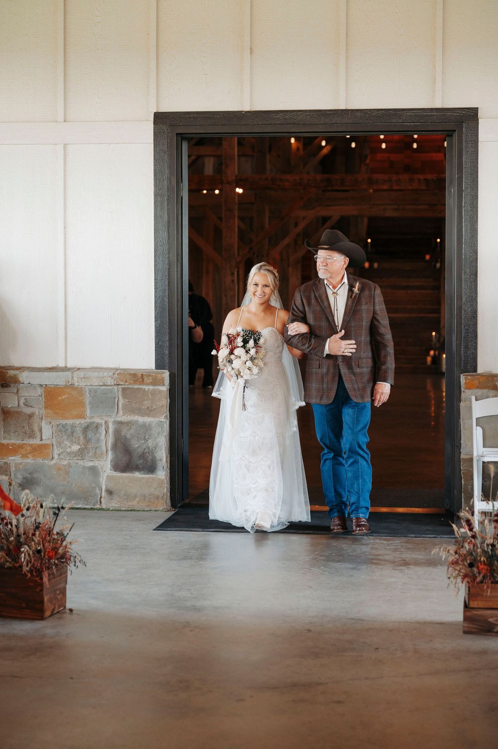 A bride in a white gown and companion exit through a rustic barn doorway decorated with potted flowers.