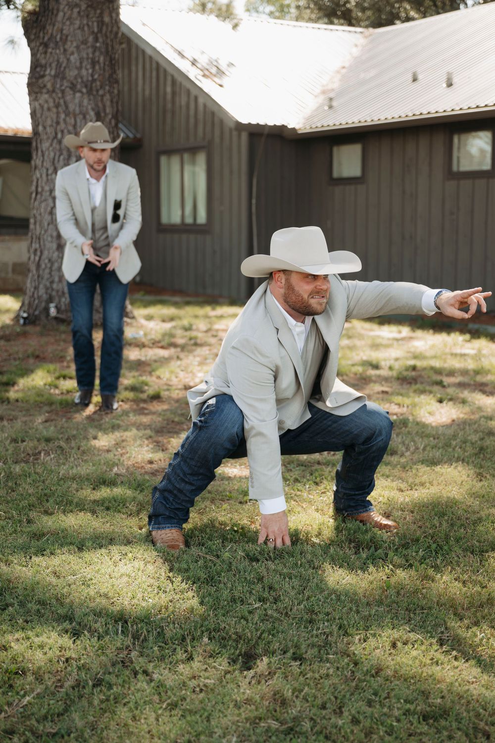 Two people in light colored jackets and cowboy hats playfully pose on a grassy lawn.