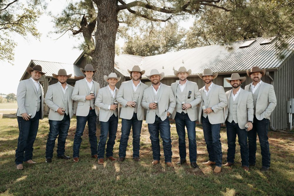 A group in matching light gray jackets and jeans stand in a line outdoors under a large tree.