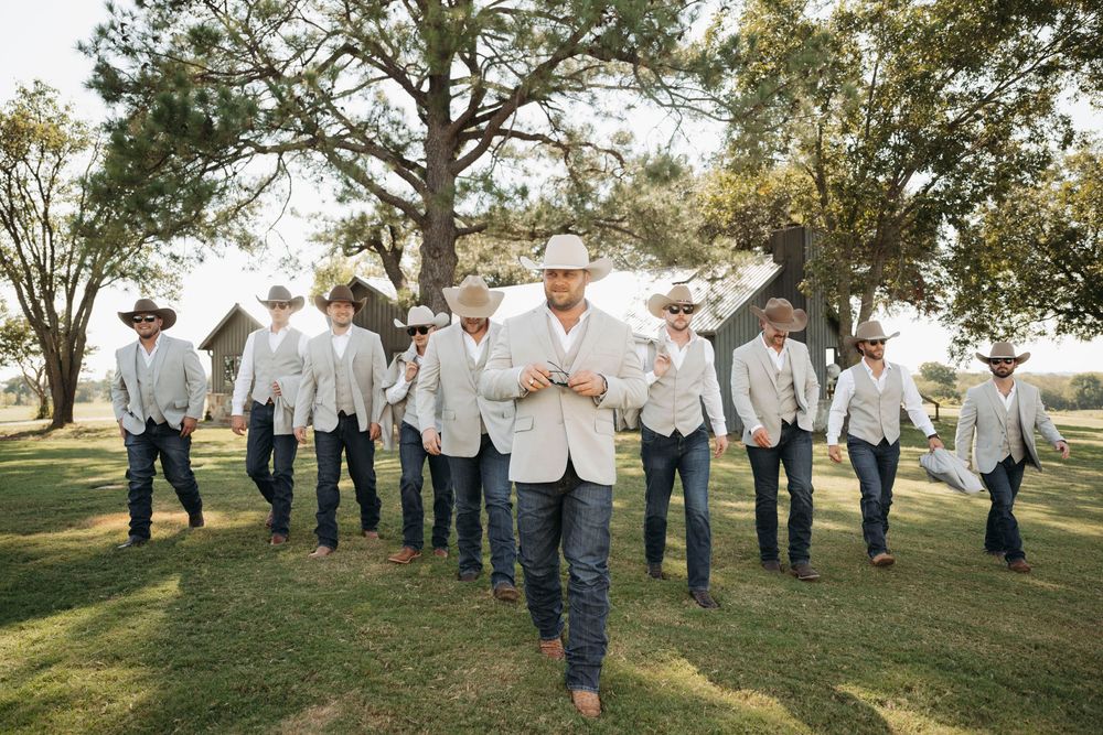 A group of groomsmen in white shirts and jeans walk together on a grassy lawn beneath large trees at an outdoor wedding.