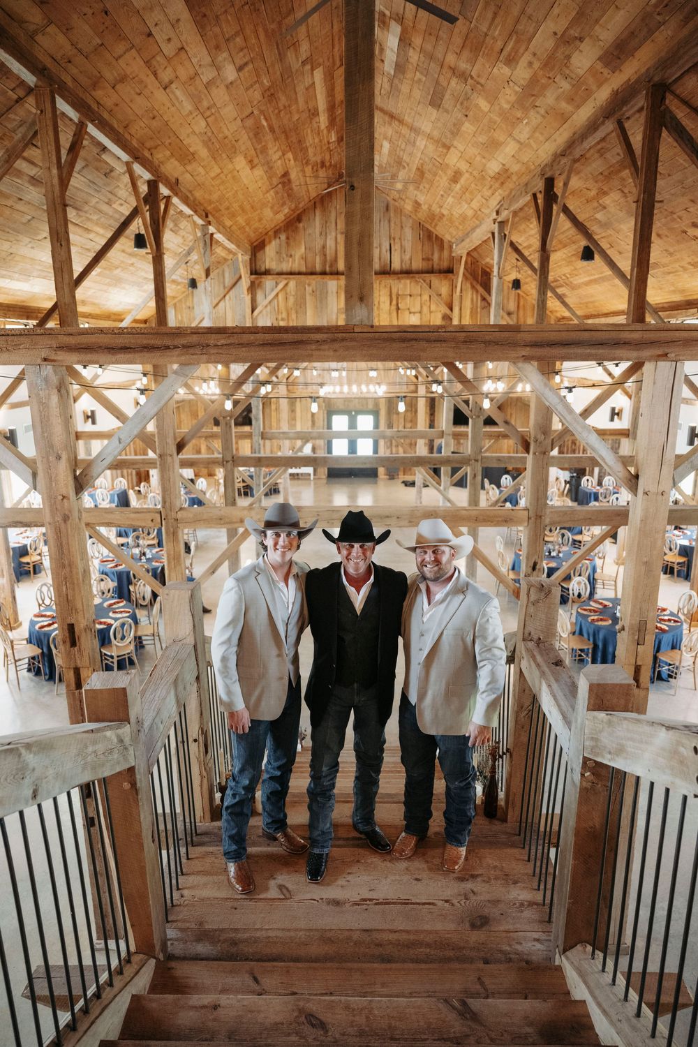 Rustic wooden barn interior with people gathered along railing in western attire at a country event.