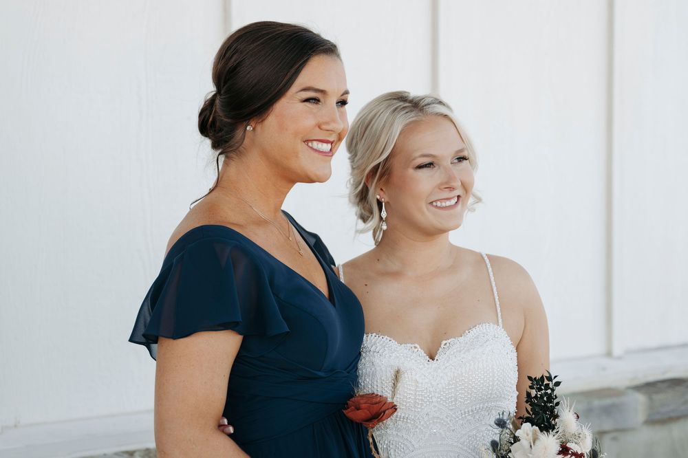 Two women in formal attire smile together at a wedding, one in navy blue and one in white.