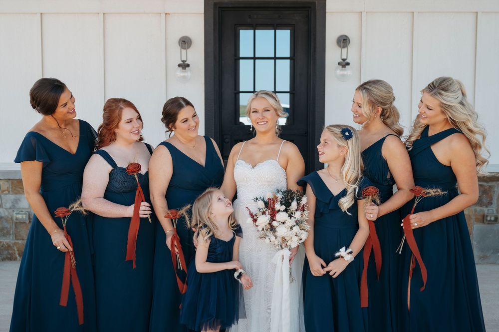 A wedding party wearing navy blue bridesmaid dresses and white bridal gown stand together against a light colored wall.