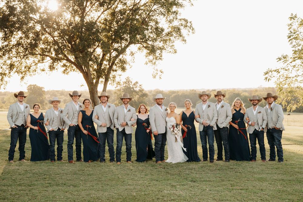 Wedding party group poses outdoors in a line against a sunset backdrop in a grassy field.