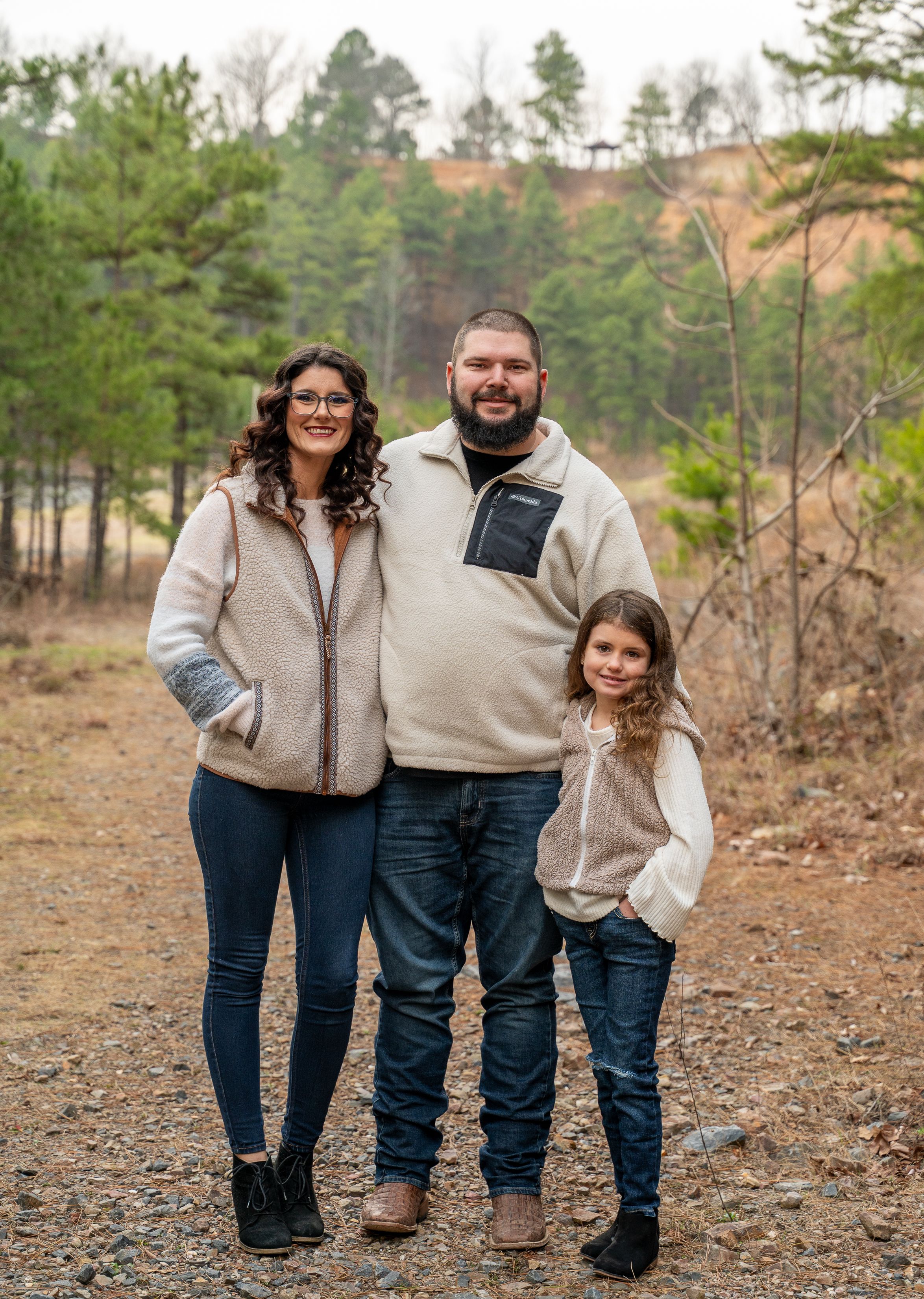 Family of three smiling together on wooded trail wearing matching cream fleece pullovers.
