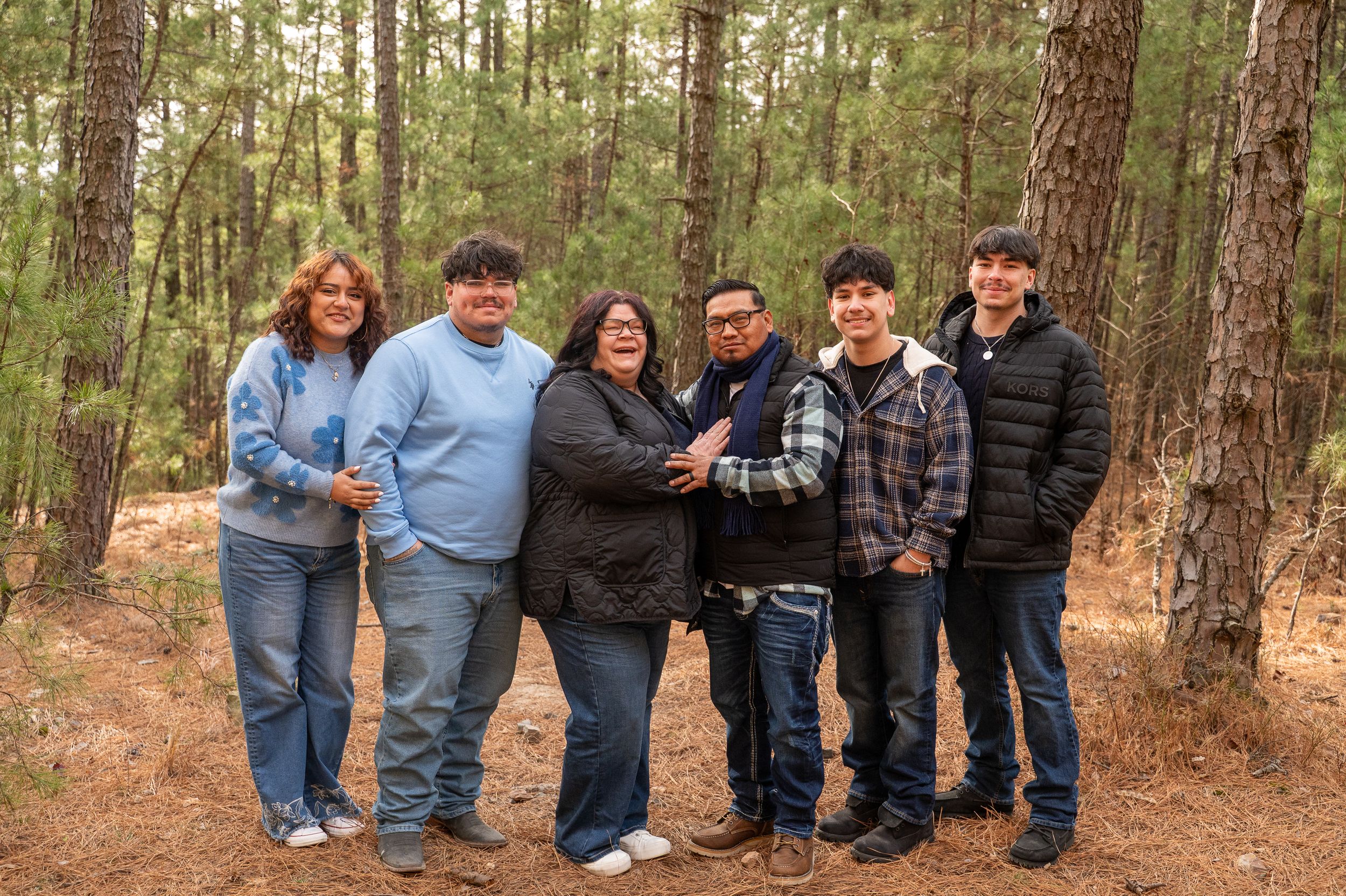 Six family members smiling together in a pine forest during an outdoor photo session.