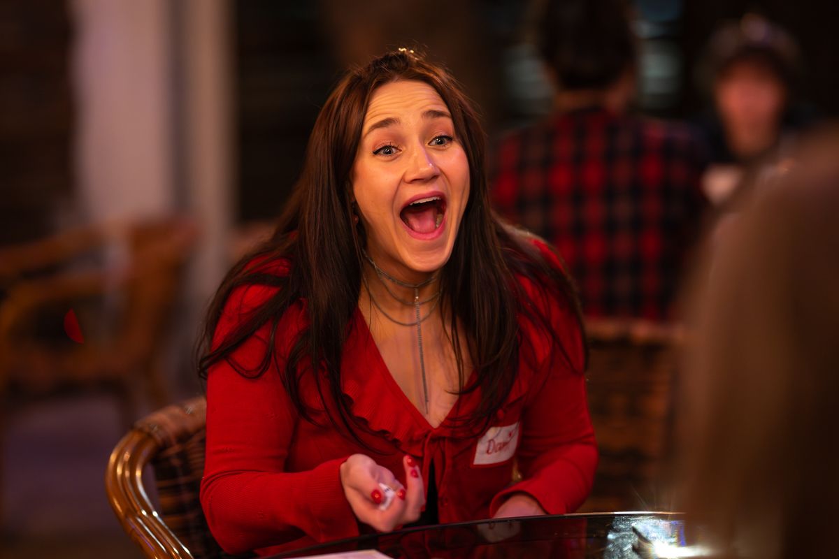 A person wearing a red sweater sits at a table expressing extreme excitement and joy with an open-mouthed expression.