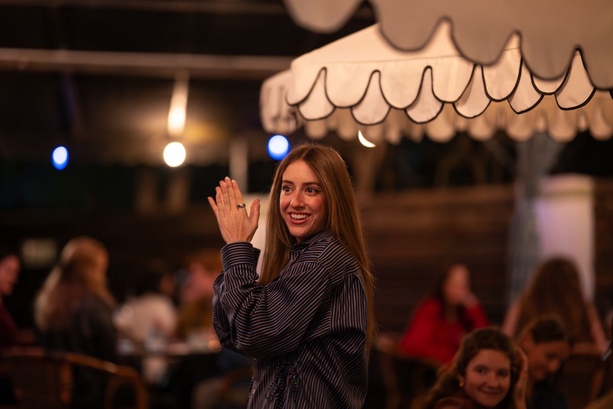 A person in a dark sweater waves at the camera while enjoying an evening at a restaurant with warm lighting and decor.