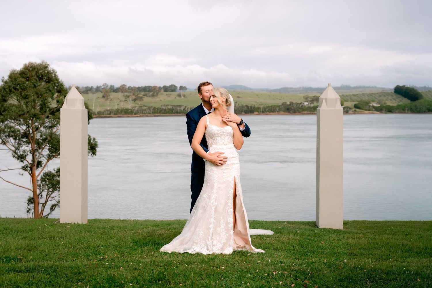 Elegant white lace wedding dress with a high slit photographed against a serene waterfront backdrop with white fence posts.