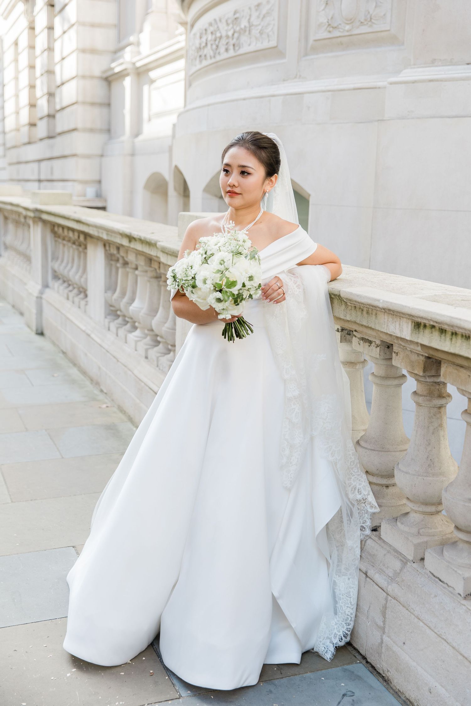 Elegant bridal portrait showcasing off-shoulder white gown and classic bouquet against ornate stone architecture outside Corinthia London