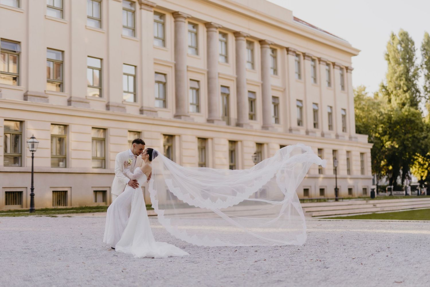 Bride in flowing white veil walking along elegant stone building exterior with classical architecture.
