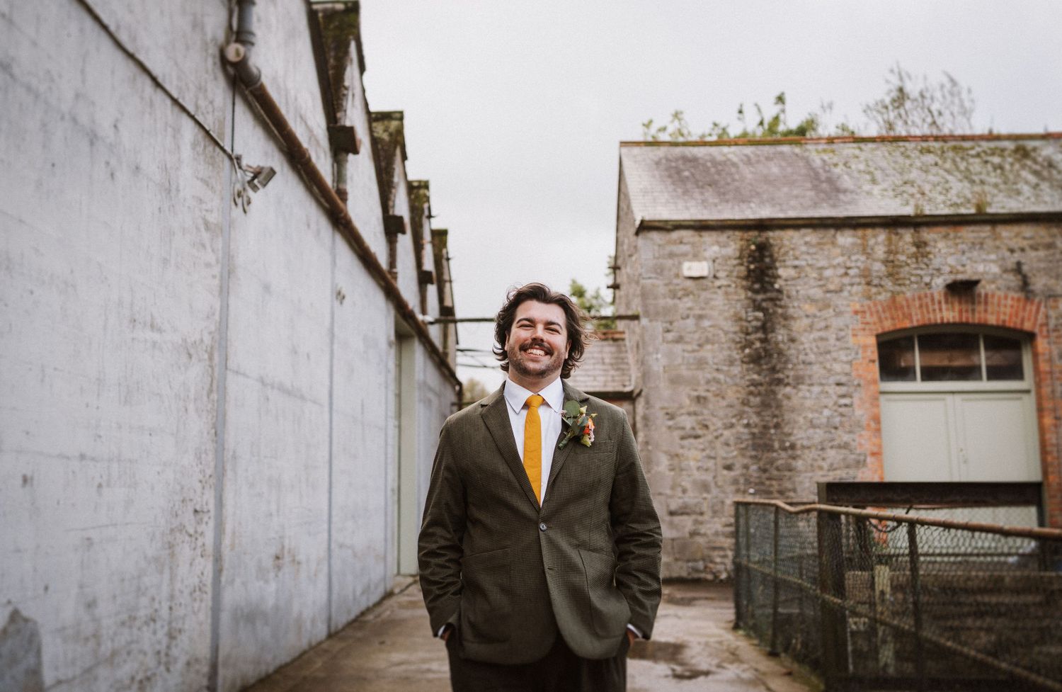 Someone in a dark suit and yellow tie stands smiling in an alleyway between concrete and stone walls.