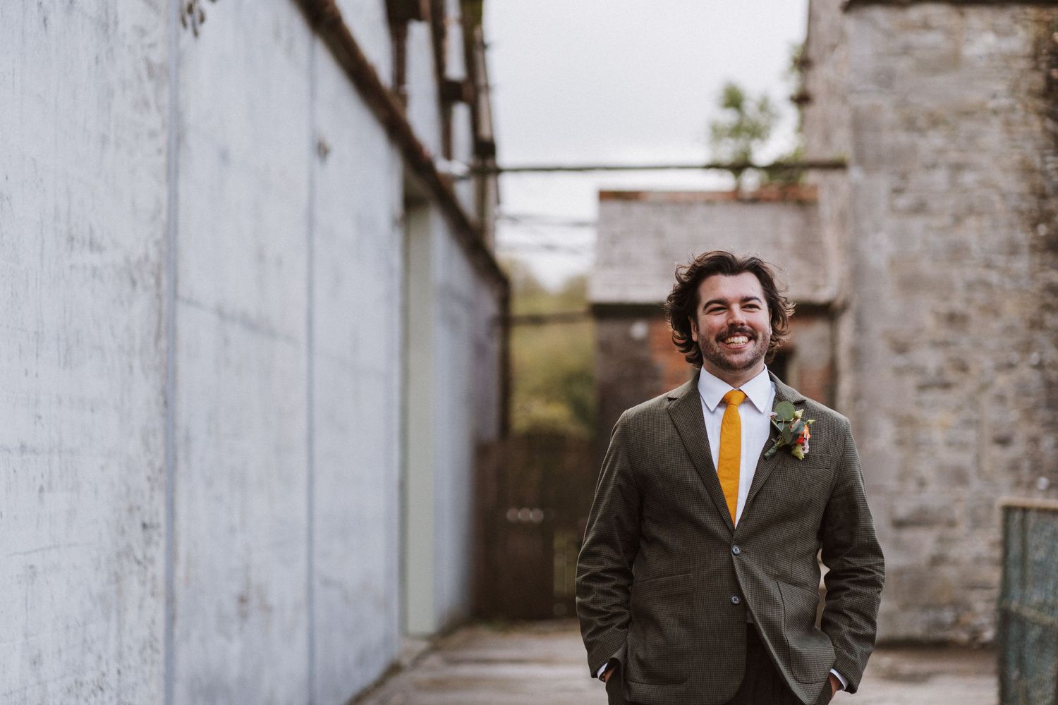 A person wearing an olive suit and orange tie poses cheerfully between industrial building walls.