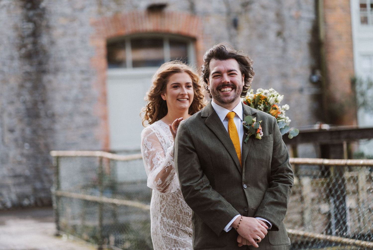 Wedding party members share candid laughs and embraces in front of a historic stone building.