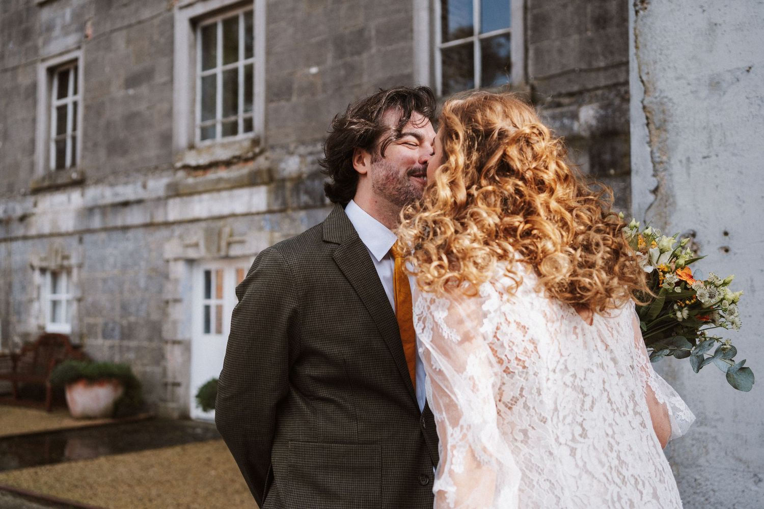 A romantic wedding couple shares tender moments in front of a stone building facade.