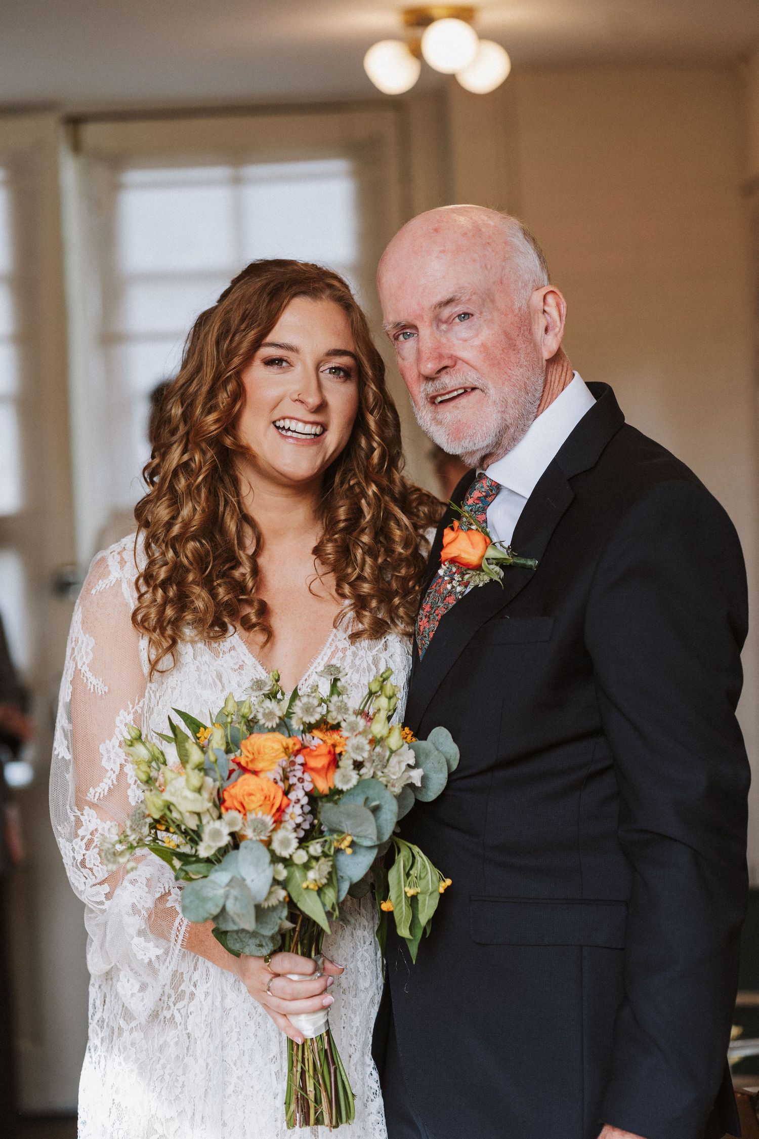 A bride in a white lace dress shares a heartwarming first look moment with an older gentleman in a black suit.