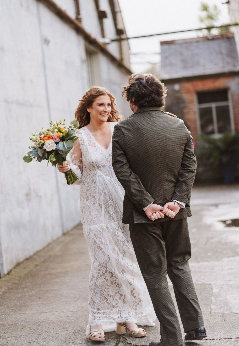A bride in a lace wedding dress holds a colorful bouquet while sharing a joyful moment with her groom in a courtyard.