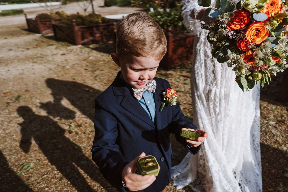 Young ring bearer in navy suit holds succulent plants at outdoor wedding ceremony with floral arrangements.