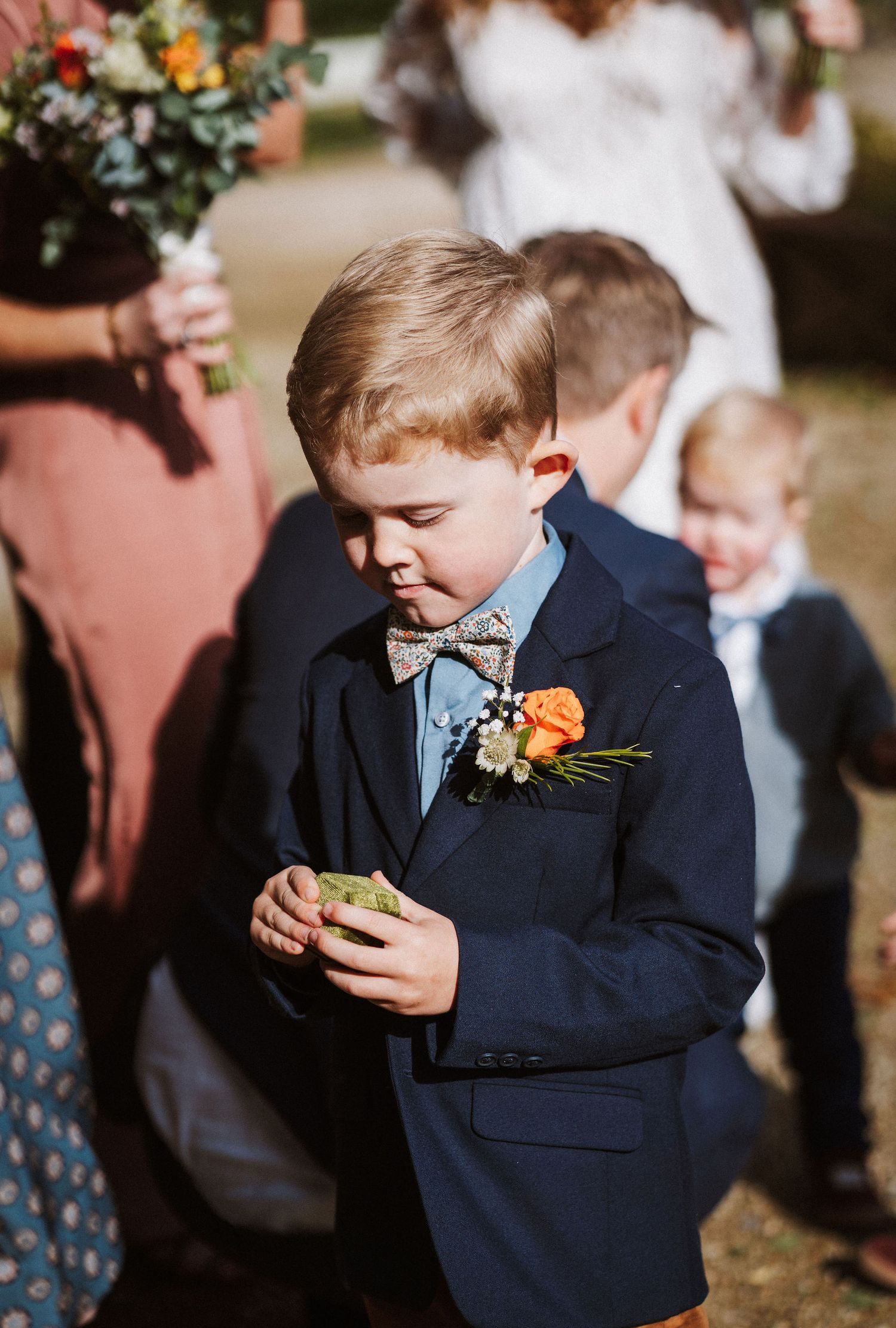 Young ring bearer in navy suit checks his phone during an outdoor wedding ceremony.